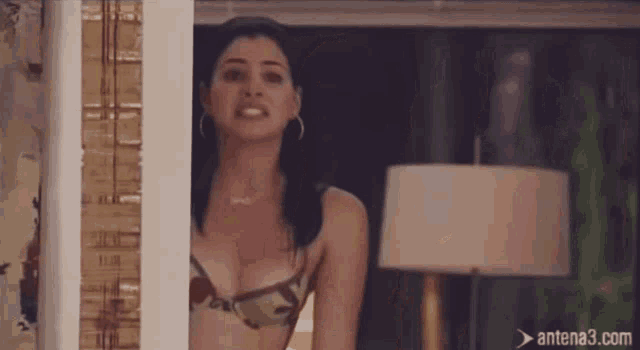 a woman in a bikini is standing in front of a window with antena3.com written on the bottom
