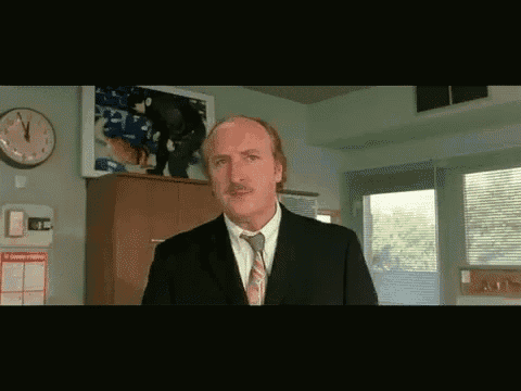 a bald man in a suit and tie is standing in front of a clock that shows the time as 12:05