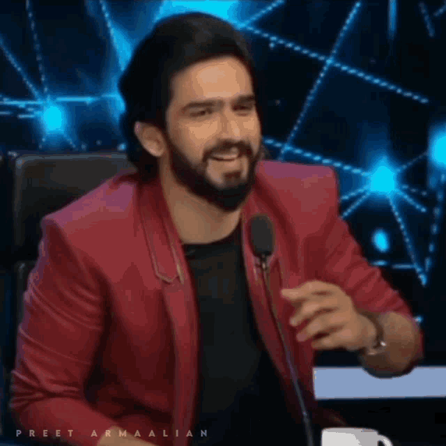 a man with a beard and a red jacket is sitting in front of a microphone .