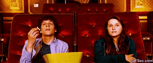 a man and a woman are sitting in a theater watching a movie and eating popcorn .