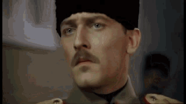 a man with a mustache wearing a hat and military uniform is looking at the camera .