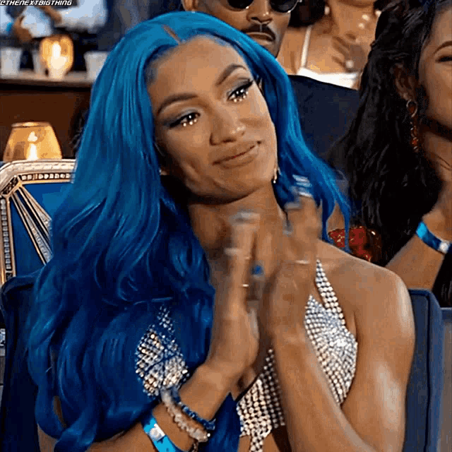 a woman with blue hair is sitting in a chair with her hands folded and smiling .