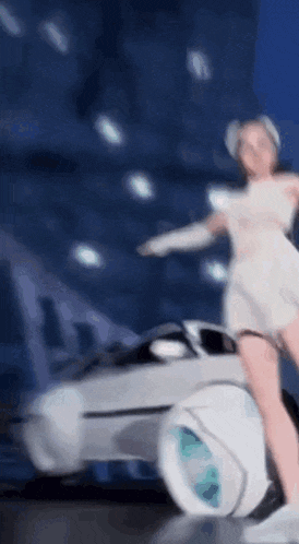 a woman in a white dress is dancing in front of a futuristic car .