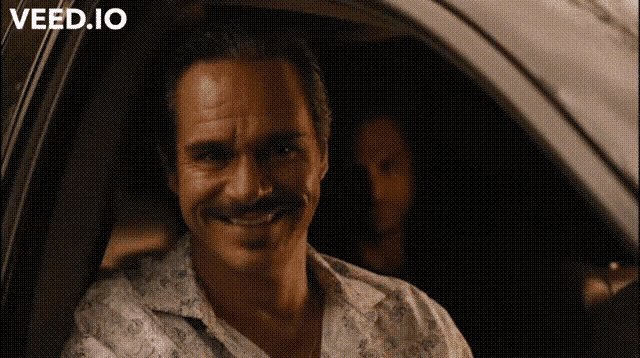 a man with a mustache is smiling while sitting in a car with the words veed.io written above him