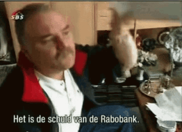 a man with a mustache is sitting in front of a table with the words het is de schuld van de rabobank on it