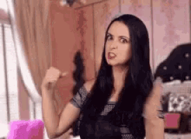 a woman with long black hair is making a funny face and pointing at herself .