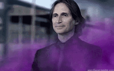 a man in a black suit and tie is standing in front of a building with purple smoke coming out of it .