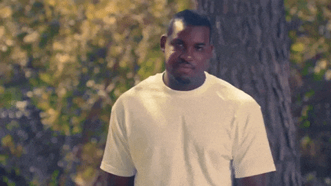 a man in a white t-shirt is standing in front of a tree and looking at the camera .
