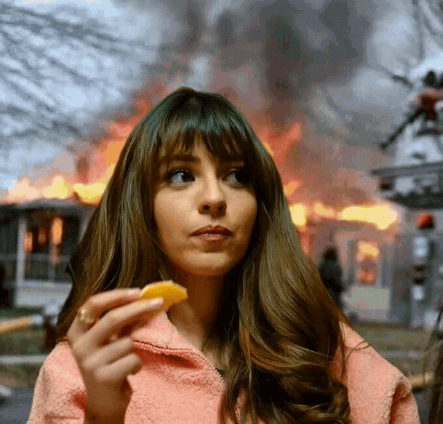 a woman in a pink jacket is holding a slice of orange in front of a burning house