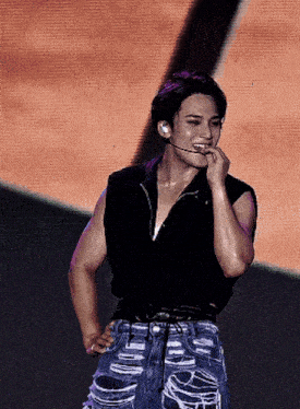 a man wearing ripped jeans and a black vest stands on a stage with his hand on his chin
