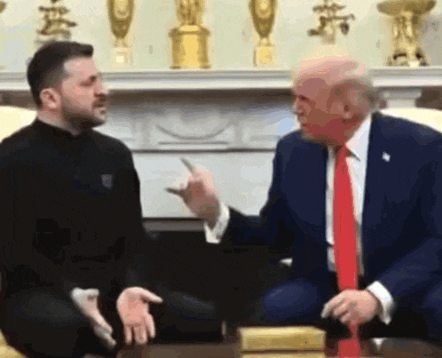 a man in a suit and tie is talking to a man in a black shirt while sitting on a couch .