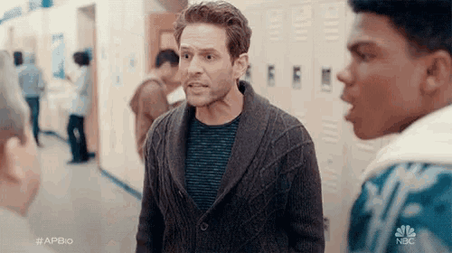 a man in a sweater is standing in a hallway talking to a group of people .