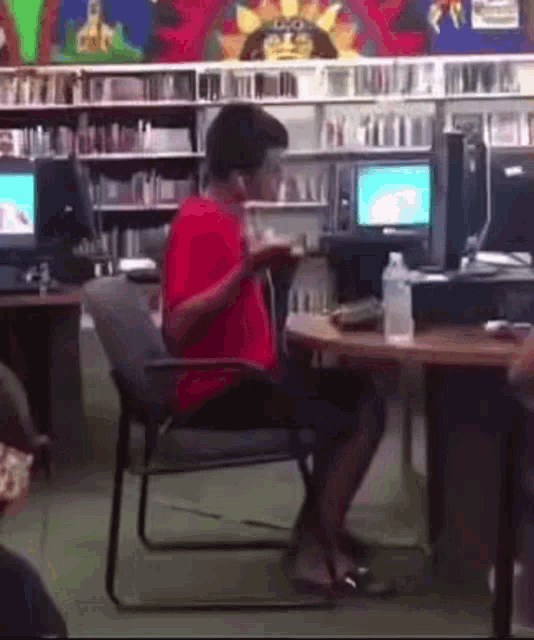 a man in a red shirt is sitting at a desk in a library .