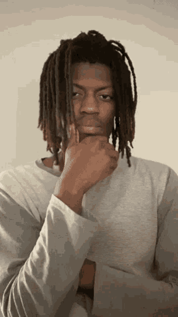 a young man with dreadlocks is wearing a gray shirt and holding his hand to his chin .