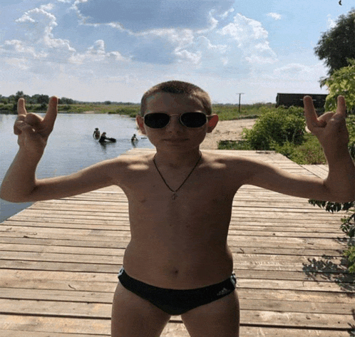 a shirtless boy wearing sunglasses stands on a wooden pier