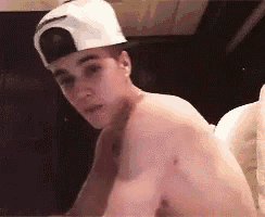 a shirtless young man wearing a baseball cap is sitting on a bed .