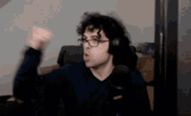 a man wearing headphones and glasses is sitting in front of a microphone with his fist in the air .