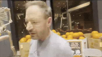 a man standing in front of a pile of oranges and a skeleton in the background