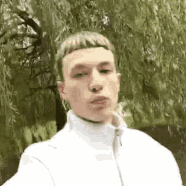 a young man wearing a white jacket is standing in front of a tree .