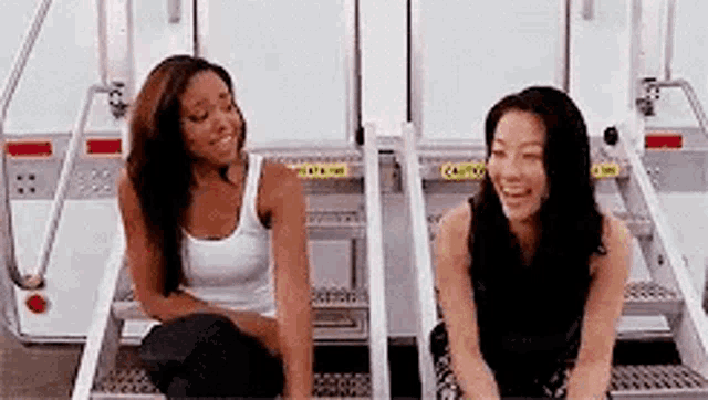 two women are sitting on a set of stairs talking to each other and smiling .