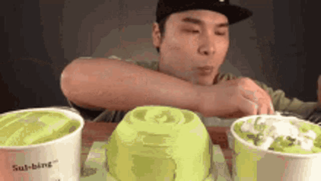 a man in a hat is sitting at a table with three cups of green ice cream .