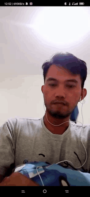 a man wearing ear buds and a grey shirt has the time of 12:52 on his phone screen