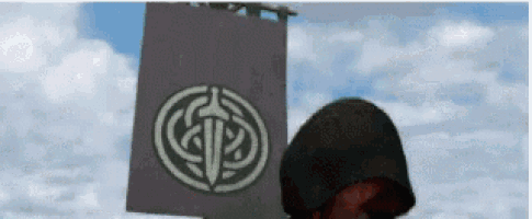 a person in a hood is standing in front of a sign with a celtic symbol on it .