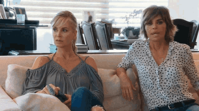 two women are sitting on a couch and one is holding a remote