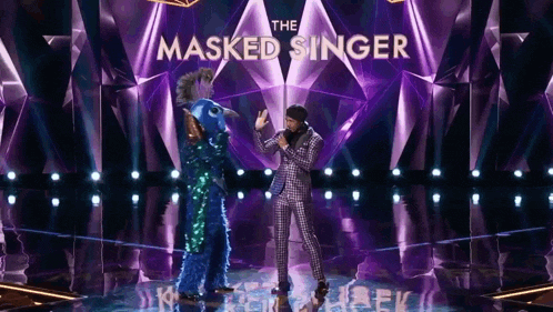 a man in a suit stands on a stage with a masked singer behind him