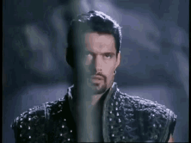 a man with a beard and piercings is standing in a dark room behind bars .