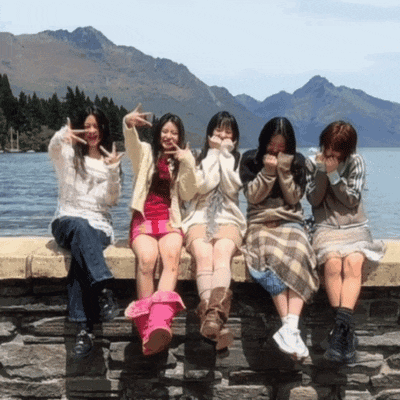 four girls sit on a stone wall with mountains in the background making peace signs
