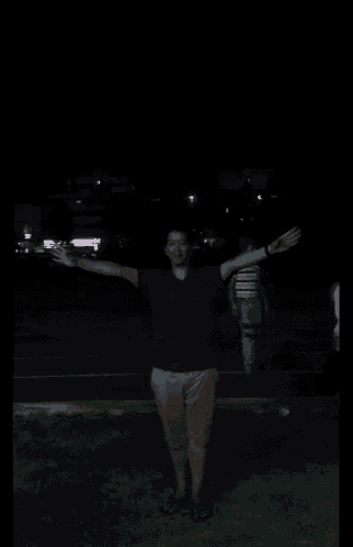 a man in a green shirt and khaki shorts stands in the dark with his arms outstretched