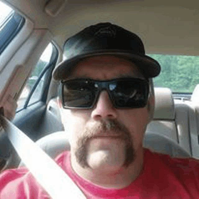 a man with a mustache and sunglasses is sitting in a car wearing a hat .