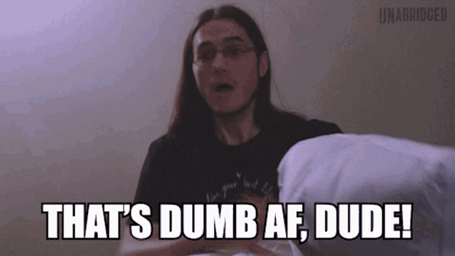 a man with long hair is holding a pillow and says that 's dumb af dude !