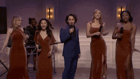 a man in a blue suit is singing into a microphone while a group of women in red dresses are dancing around him