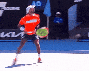 a man in an orange shirt is holding a tennis racquet in front of a kia sign
