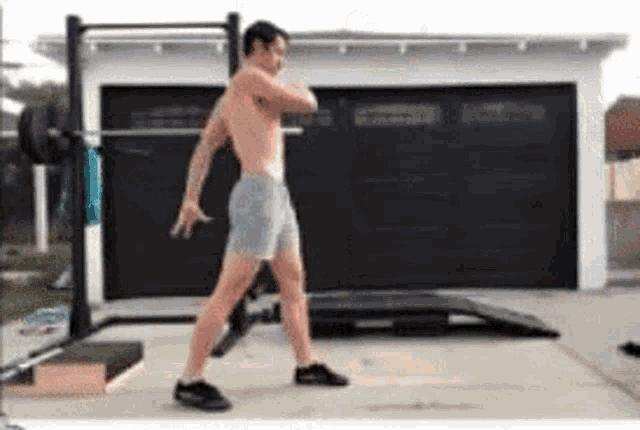 a shirtless man is standing in front of a garage with a barbell in his hands .