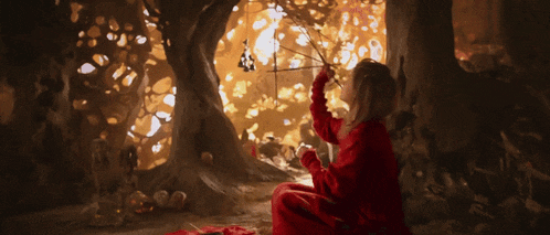 a little girl in a red dress is sitting under a tree holding a stick
