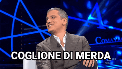a man in a suit is sitting in front of a blue background that says coglione di merda