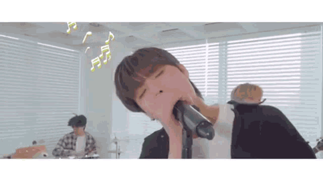 a young man is singing into a microphone in a room with a drummer in the background .
