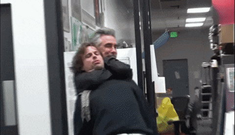 a man is carrying another man on his back in a room with a green exit sign hanging from the ceiling