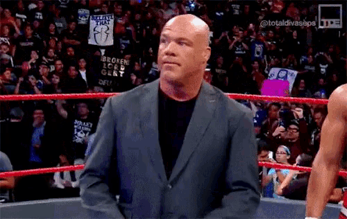 a bald man in a suit is standing in a wrestling ring with a crowd watching .