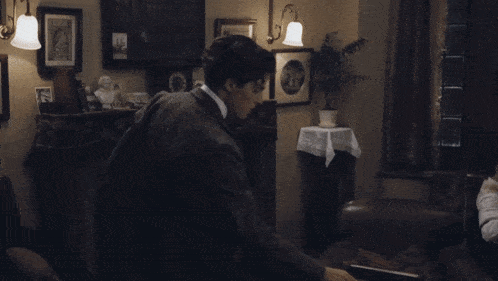 a man in a suit stands in a living room looking at a book