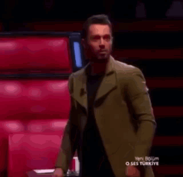 a man in a green jacket and black shirt is standing in front of a red chair .