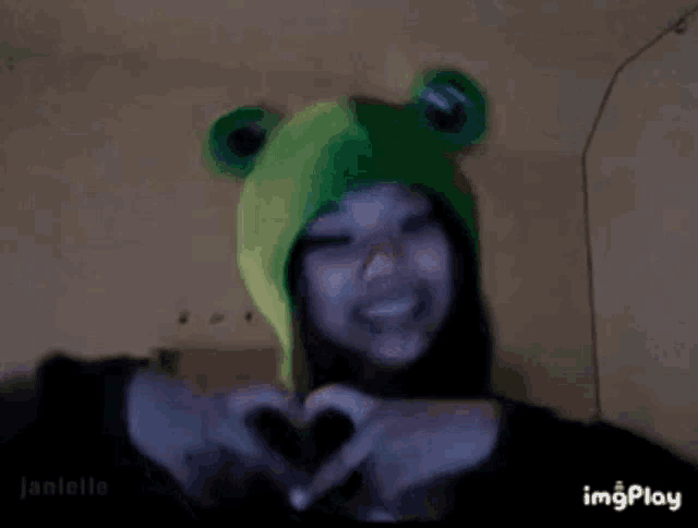 a girl wearing a frog hat is making a heart with her hands .