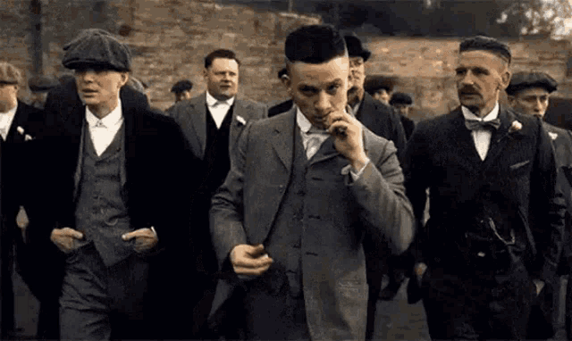 a group of men in suits and hats are walking down a street . one of the men is smoking a cigarette .