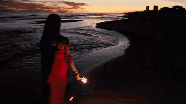 a woman in a red dress is walking on the beach at night holding a sparkler