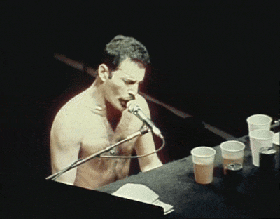 a shirtless man sings into a microphone while playing a keyboard