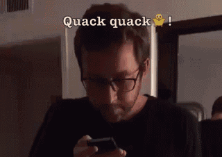 a man wearing glasses is looking at his cell phone with the words quack quack behind him