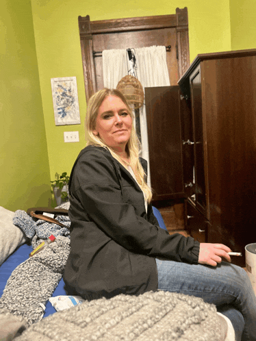 a woman in a black jacket is sitting on a bed smoking a cigarette
